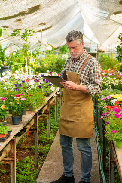 A 55-year-old worker in a vivid plant nursery, documenting his tasks and customer inquiries in a notebook. - Foto, Imagem