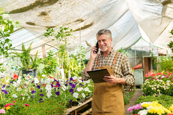 Dedykowany pracownik szkółki kwiatowej wielozadaniowy, z uwagą przyjmując zamówienie kwiatowe klienta podczas rozmowy telefonicznej, trzymając folder. - Zdjęcie, obraz