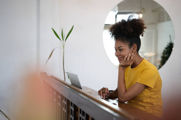 幸せな若いアフリカ系アメリカ人の女性を使用してノートパソコン,コーヒーショップで木製のカウンターに座ってリモートで作業.  - 写真・画像