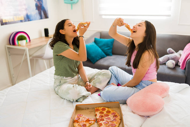 Happy teenage girls enjoying eating delicious pizza together at home while having fun hanging out in bed  - Photo, Image