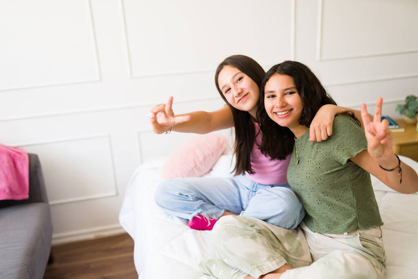 Ευτυχισμένες έφηβες κοπέλες που κάνουν το σήμα ειρήνης και χαμογελούν διασκεδάζοντας μαζί στην κρεβατοκάμαρα ενώ κάνουν οπτική επαφή  - Φωτογραφία, εικόνα