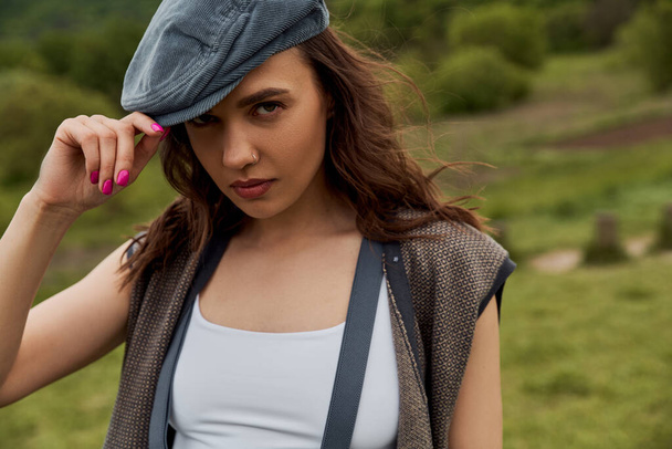 Portrait of stylish brunette woman in vintage clothes, vest and suspenders touching newsboy cap and looking at camera with blurred landscape at background, fashion-forward in countryside - Photo, Image