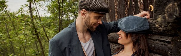 Smiling and fashionable man in jacket and newsboy cap looking at cheerful girlfriend while standing together near rustic house at nature, stylish couple in rural setting, banner  - Photo, Image