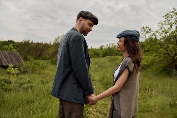 Bearded and stylish man in jacket and newsboy cap holding hand and looking at cheerful girlfriend in vest and standing together on grassy lawn at overcast, stylish couple in rural setting - Photo, Image