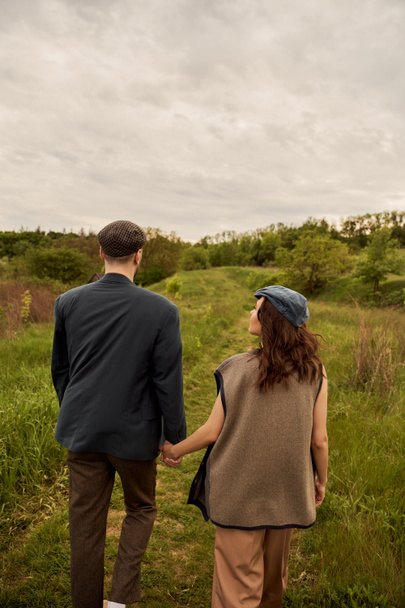 Brunette et femme à la mode en gilet et bonnet de newsboy tenant la main du petit ami en veste tout en marchant ensemble sur la prairie avec la nature et le ciel en arrière-plan, avant-garde de la mode dans la campagne - Photo, image