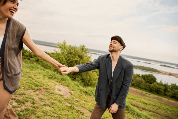 Cheerful and stylish woman in vest holding hand of bearded boyfriend in newsboy cap and jeans with scenic nature and overcast sky at background, fashion-forwards in countryside - Photo, Image