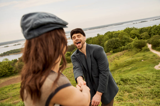 Veselý a trendy vousatý muž v bundě a novinářská čepice drží ruku rozmazané přítelkyně, zatímco stojí s malebnou krajinou a oblohou na pozadí, módní-vpřed na venkově - Fotografie, Obrázek