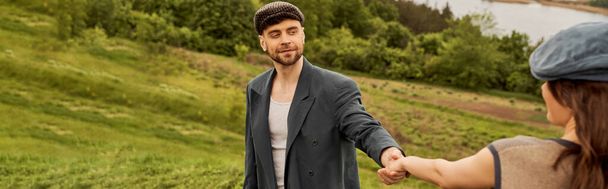 Smiling and fashionable bearded man in newsboy cap and jacket holding hand of blurred brunette girlfriend while spending time with nature at background, fashionable couple in countryside, banner  - Photo, Image