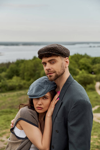 Portrait of confident bearded man in jacket hugging brunette girlfriend in newsboy cap and vest and looking at camera while standing with landscape at background, fashionable couple in countryside - Photo, Image