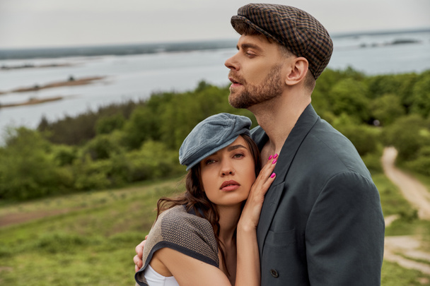 Brunette and fashionable woman in newsboy cap and vest hugging bearded boyfriend in jacket and standing with scenic landscape at background, fashionable couple in countryside - Photo, Image