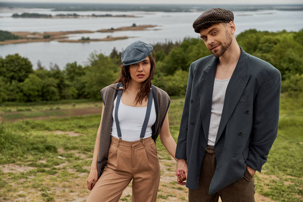 Confident and bearded man in jacket holding hand of fashionable girlfriend in newsboy cap and suspenders and looking at camera while standing with nature at background, stylish pair amidst nature - Photo, Image