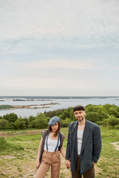 Smiling bearded man in vintage-inspired outfit holding hand of brunette girlfriend in suspenders and newsboy cap and standing with nature at background, stylish pair amidst nature - Fotografie, Obrázek