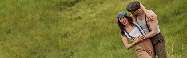 Trendy bearded man in vintage outfit and sunglasses hugging positive girlfriend in newsboy cap and standing on blurred grassy lawn at background, stylish pair amidst nature, banner  - Photo, Image