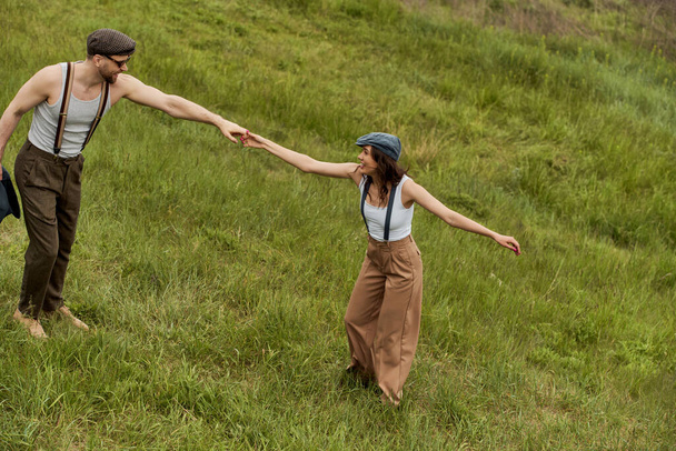 Side view of positive and fashionable couple in newsboy caps and suspenders holding hands and having fun on grassy meadow at background, stylish couple enjoying country life - Φωτογραφία, εικόνα
