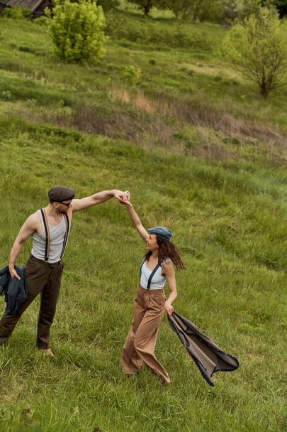 Side view of cheerful brunette woman in vintage clothes holding vest and having fun with boyfriend in newsboy cap and sunglasses on grassy meadow, stylish couple enjoying country life - Photo, Image