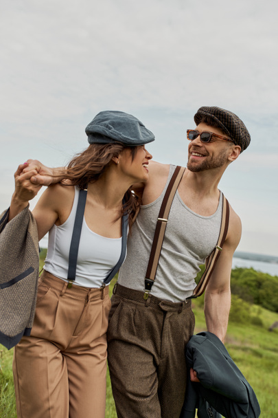 Lächelnder bärtiger Mann im Vintage-Outfit und Kioskmütze umarmt brünette Freundin mit Hosenträgern und spaziert vor malerischer Landschaft im Hintergrund, stilvolles Paar genießt das Landleben - Foto, Bild