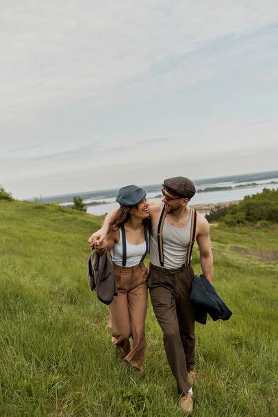 Cheerful and fashionable romantic couple in newsboy caps, vintage outfits and suspenders hugging and walking together on grassy hill at background, trendy couple in the rustic outdoors - Photo, Image