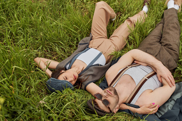 Top view of stylish brunette woman in vintage outfit and newsboy cap hugging bearded boyfriend in sunglasses while lying together and spending time on grassy meadow, stylish partners in rural escape - Foto, immagini