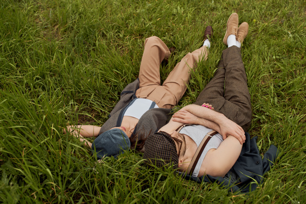 ニュースボーイキャップとヴィンテージの衣装でファッショナブルなロマンチックなカップルの高い角度ビューが横たわっています,草の牧草地で一緒に過ごす時間とリラックス,農村部のエスケープでスタイリッシュなパートナー,ロマンチックな休暇 - 写真・画像