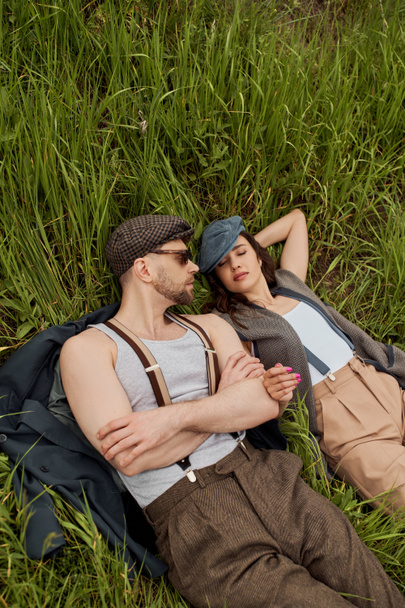 Horní pohled na vousatý muž v slunečních brýlích a suspendéry ležící na bundě v blízkosti stylové přítelkyně v vintage outfit při relaxaci na travnatém poli, módní pár obklopen přírodou - Fotografie, Obrázek