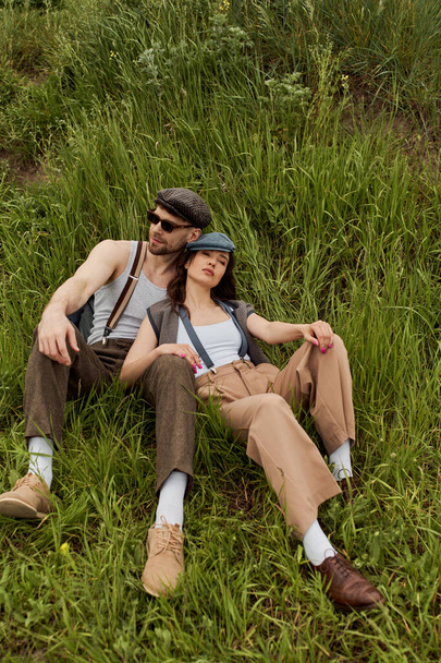 Trendi romantikus pár évjárat ruhák és újságos sapkák pihenő egymás mellett, miközben ül a dombon zöld fű nyáron, divatos pár körül a természet, romantikus menekülés - Fotó, kép