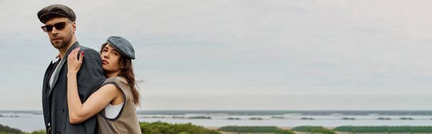 Stylowa brunetka w kamizelce i vintage strój obejmujący chłopaka w kurtce i okularach przeciwsłonecznych patrząc na aparat fotograficzny z krajobrazem w tle, modne dwa w rustykalnym otoczeniu, baner  - Zdjęcie, obraz