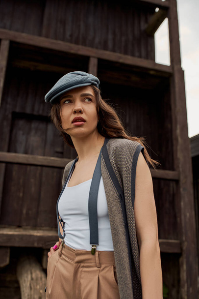 Portrait of fashionable brunette woman in newsboy cap, suspenders and vest holding hand in pocket of pants and looking away near rustic house in rural setting, vintage-inspired clothing - Photo, Image