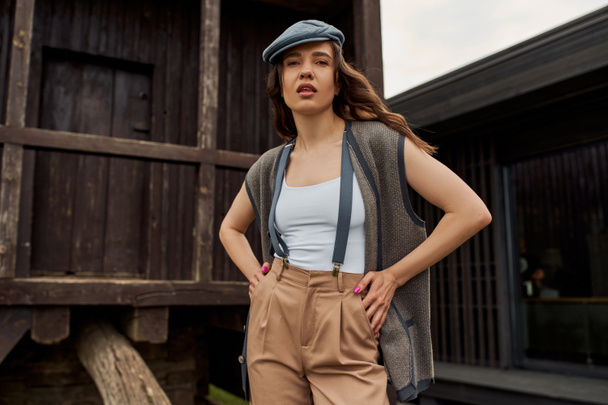 Κομψό μελαχρινή γυναίκα σε vintage ντύσιμο και newsboy καπέλο ποζάρουν σε γιλέκο και τιράντες, ενώ στέκεται κοντά ρουστίκ σπίτι σε αγροτικό περιβάλλον το καλοκαίρι, vintage-inspired ρούχα - Φωτογραφία, εικόνα