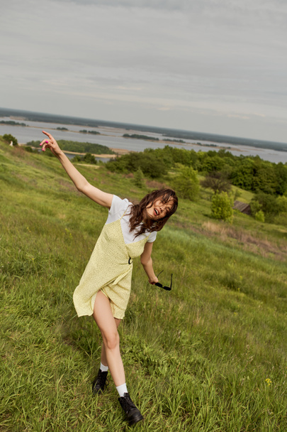 Mujer morena con estilo en vestido de sol y botas de moda sosteniendo gafas de sol mientras se divierten y de pie en la hierba con paisaje escénico en el fondo en verano, alegría de verano - Foto, Imagen