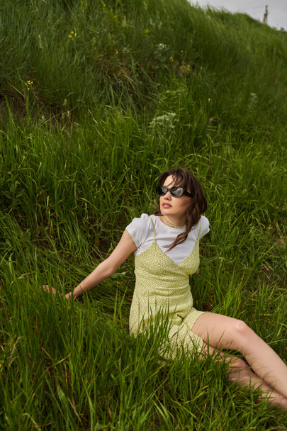 Femme brune élégante en lunettes de soleil et robe de soleil se détendre et passer du temps assis sur la colline avec de l'herbe verte à l'arrière-plan, paysage naturel et concept libre d'esprit - Photo, image
