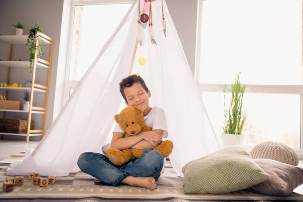 Portrait of adorable small schoolboy embrace favorite plushie toy wear white shirt modern interior living room playroom daycare indoors. - 写真・画像