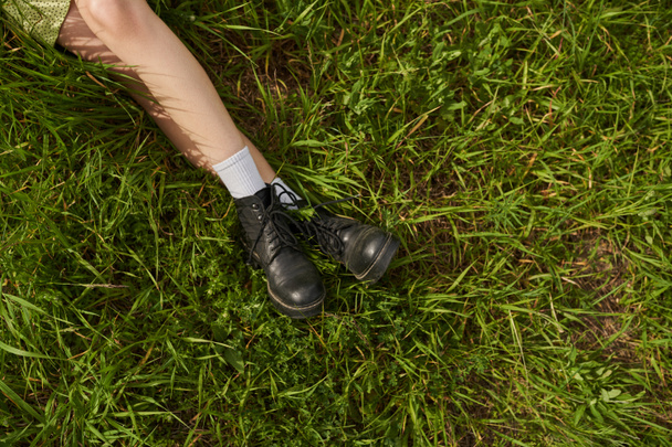 Blick von oben auf die Beine einer stilvollen Frau in Stiefeln, die auf einer Wiese mit grünem Gras, natürlicher Landschaft und freigeistigem Konzept sitzt und relaxt - Foto, Bild