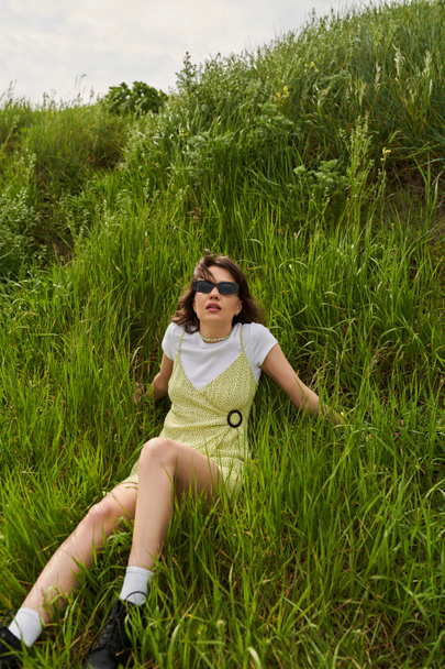 Fashionable brunette woman in sunglasses and sundress spending time and relaxing on green meadow with grass, peaceful retreat and relaxing in nature concept, stylish woman - Photo, Image
