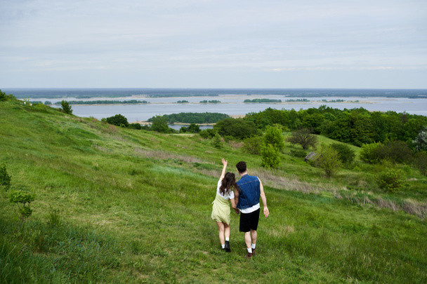 Back view of stylish couple in summer outfits holding hands and standing on grassy field with scenic view and cloudy sky at background, couple in love enjoying nature, tranquility - Photo, Image