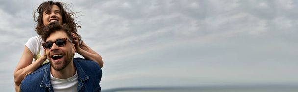 Positive brünette Freundin im Sommeroutfit huckepack auf stylischem Freund in Sonnenbrille und Jeansweste im Stehen mit wolkenverhangenem Himmel im Hintergrund, Abenteuer auf dem Land und Liebesgeschichte, Banner - Foto, Bild