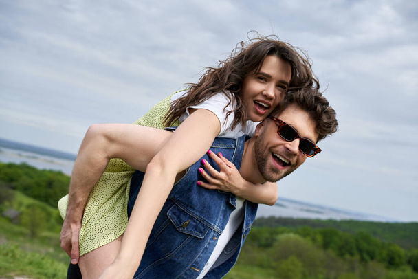 Excited stylish woman in sundress piggybacking on cheerful boyfriend in denim vest and sunglasses while spending time on nature, countryside adventure and love story, tranquility - Zdjęcie, obraz