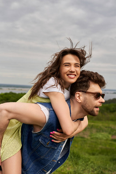 Joyful brunette woman in sundress hugging bearded boyfriend in sunglasses and denim vest and having fun on nature at background, countryside adventure and love story, tranquility - Photo, Image