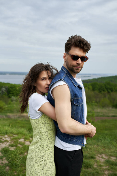 Stylish woman in sundress embracing bearded boyfriend in sunglasses and denim vest and looking at camera while standing with landscape and sky at background, love story and countryside adventure - Photo, Image
