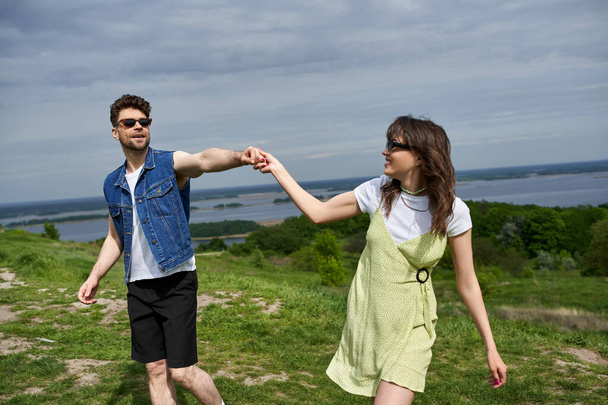 Smiling brunette man in sunglasses and denim vest holding hand of stylish girlfriend in sundress and spending time on grassy hill with cloudy sky at background, countryside wanderlust and love concept - Photo, Image