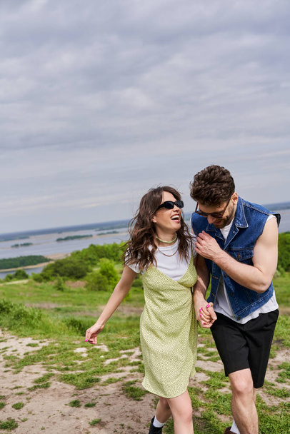 Cheerful brunette man in stylish summer outfit and denim vest holding hand of excited girlfriend in sunglasses and sundress and walking together on path on grassy hill, countryside leisurely stroll - Фото, изображение
