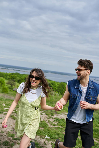 Positive brünette Mann mit Sonnenbrille und Jeansweste hält Hand der fröhlichen Freundin in stilvoller Kleidung und Zeit mit der Natur im Hintergrund verbringen, ländliche gemütliche Spaziergänge, Ruhe - Foto, Bild