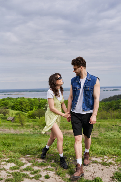Cheerful and stylish brunette woman in sundress and boots holding hand and talking to bearded boyfriend in denim vest and sunglasses while standing in nature, countryside leisurely stroll - Photo, Image