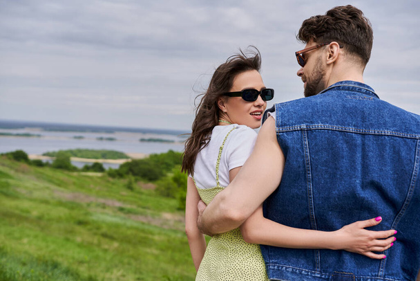 Fröhliche und modische brünette Frau mit Sonnenbrille und Sonnenbrille, die ihren bärtigen Freund in Jeansweste umarmt, während sie im Hintergrund auf einem verschwommenen Grashügel steht, Konzept eines ländlichen Rückzugs - Foto, Bild
