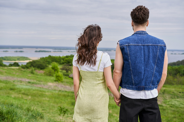 Vista trasera de la pareja romántica con estilo en trajes de verano tomados de la mano mientras están de pie juntos en la colina cubierta de hierba con paisaje panorámico borroso en el fondo, concepto de retiro rural - Foto, Imagen