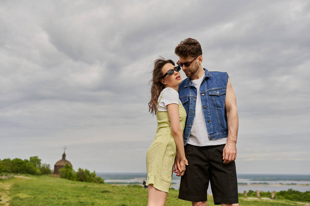 Stilvoller bärtiger Mann in Jeansweste, der die Hand einer romantischen Freundin mit Sonnenbrille und Sonnenbrille berührt, während er im Hintergrund vor ländlicher Kulisse steht, ländliches Rückzugskonzept - Foto, Bild