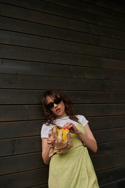 Mujer morena de moda en gafas de sol y traje de verano celebración de pan fresco en paquete de artesanía, mientras que de pie cerca de la casa de madera en el fondo, concepto de vibraciones de verano, tranquilidad - Foto, Imagen