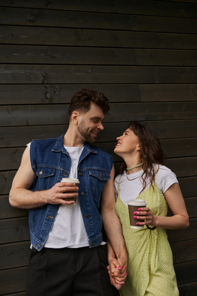 Cheerful and stylish romantic couple in summer outfits looking at each other and holding coffee to go while standing near wooden house in rural setting, carefree moments concept - Photo, Image