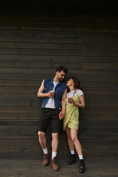 Πλήρες μήκος της μόδας ρομαντικό ζευγάρι σε μπότες και καλοκαιρινά ρούχα κρατώντας καφέ για να πάει και κοιτάζοντας ο ένας τον άλλο, ενώ στέκεται κοντά στο ξύλινο σπίτι, ανέμελες στιγμές έννοια - Φωτογραφία, εικόνα