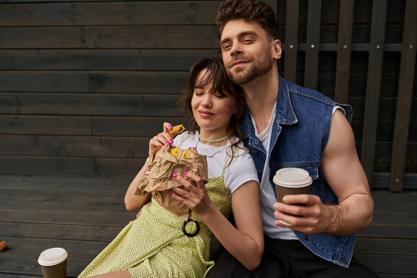 Pozitivní vousatý muž v džínové vestě drží kávu jít a sedí v blízkosti stylové brunetky přítelkyně v Sundress drží chutnou housku a dřevěný dům na pozadí, klidné prostředí koncepce - Fotografie, Obrázek