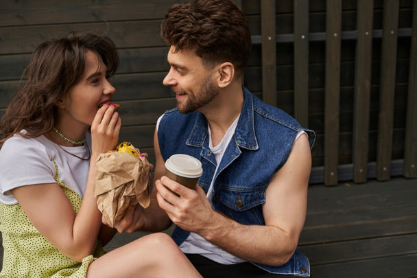 Smiling bearded man in denim vest holding coffee to go and talking to girlfriend in summer outfit eating fresh bun and sitting near wooden house at background, serene ambiance concept - Fotoğraf, Görsel